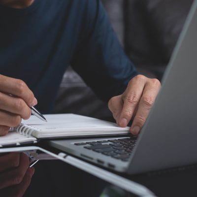 Casual business man sitting at wooden table writing note on paper notebook while working on laptop computer in office. Student attending online course and doing homework. Online education,e-learning concept